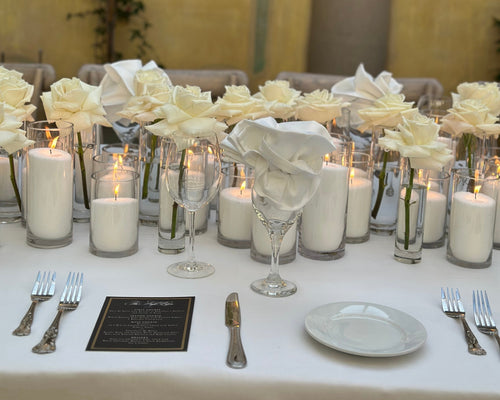 Elegant table setting with white flowers, candles, and silverware against a decorative wall.