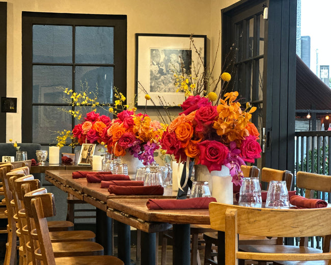 Dining room with wooden tables and chairs, colorful flower arrangements, and a painting on the wall.